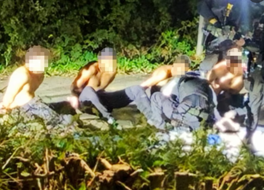Image shows shirtless men being arrested in a dark country lane. Their faces are blurred