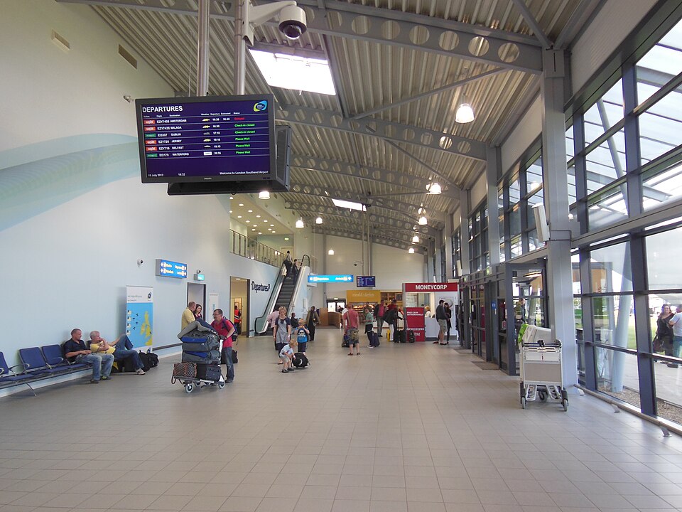 Image shows the inside of London Southend Airport terminal building during the day in 2012. A departures board is visible as well as passengers with luggage and trolleys in the background