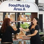 A traveler picks up a brown paper food bag from an OTG staff member at a United Airlines "Food Pick Up Area A" station inside an airport terminal.