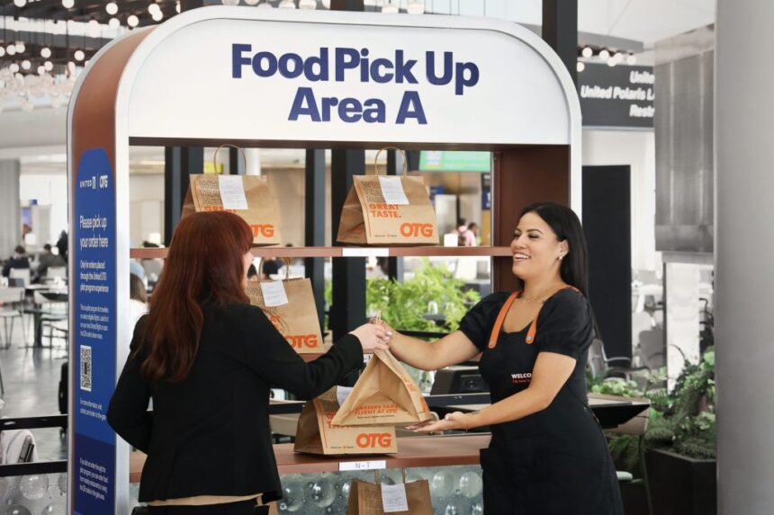 A traveler picks up a brown paper food bag from an OTG staff member at a United Airlines "Food Pick Up Area A" station inside an airport terminal.