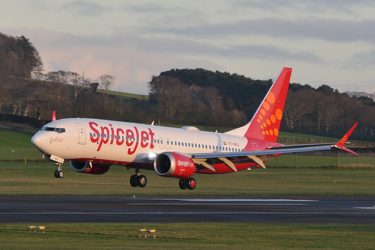 A SpiceJet Boeing 738 aircraft landing on a runway. The plane has SpiceJet's usual colours, bright red and orange, with a dotted design on the plane's tail. The sky is cloudy, and the sun is out of view.