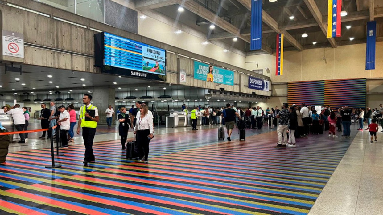 An internal view of the bustling terminal of Simón Bolívar International Airport, featuring a large, vibrant, multi-colored floor pattern that extends through the main thoroughfare. Travelers, security personnel, and airline staff move along the floor, flanked by airport infrastructure like a digital flight information display and various signs. Large vertical blue, yellow, and red banners with stars hang from the ceiling.
