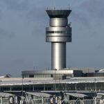An image of an air traffic control tower peaking over the top of the airport exterior. The background is a blue/grey sky.