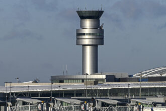 An image of an air traffic control tower peaking over the top of the airport exterior. The background is a blue/grey sky.