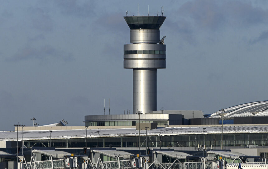An image of an air traffic control tower peaking over the top of the airport exterior. The background is a blue/grey sky.