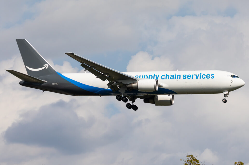 An Amazon Air cargo plane mid-flight, with a slightly cloudy sky as the background.