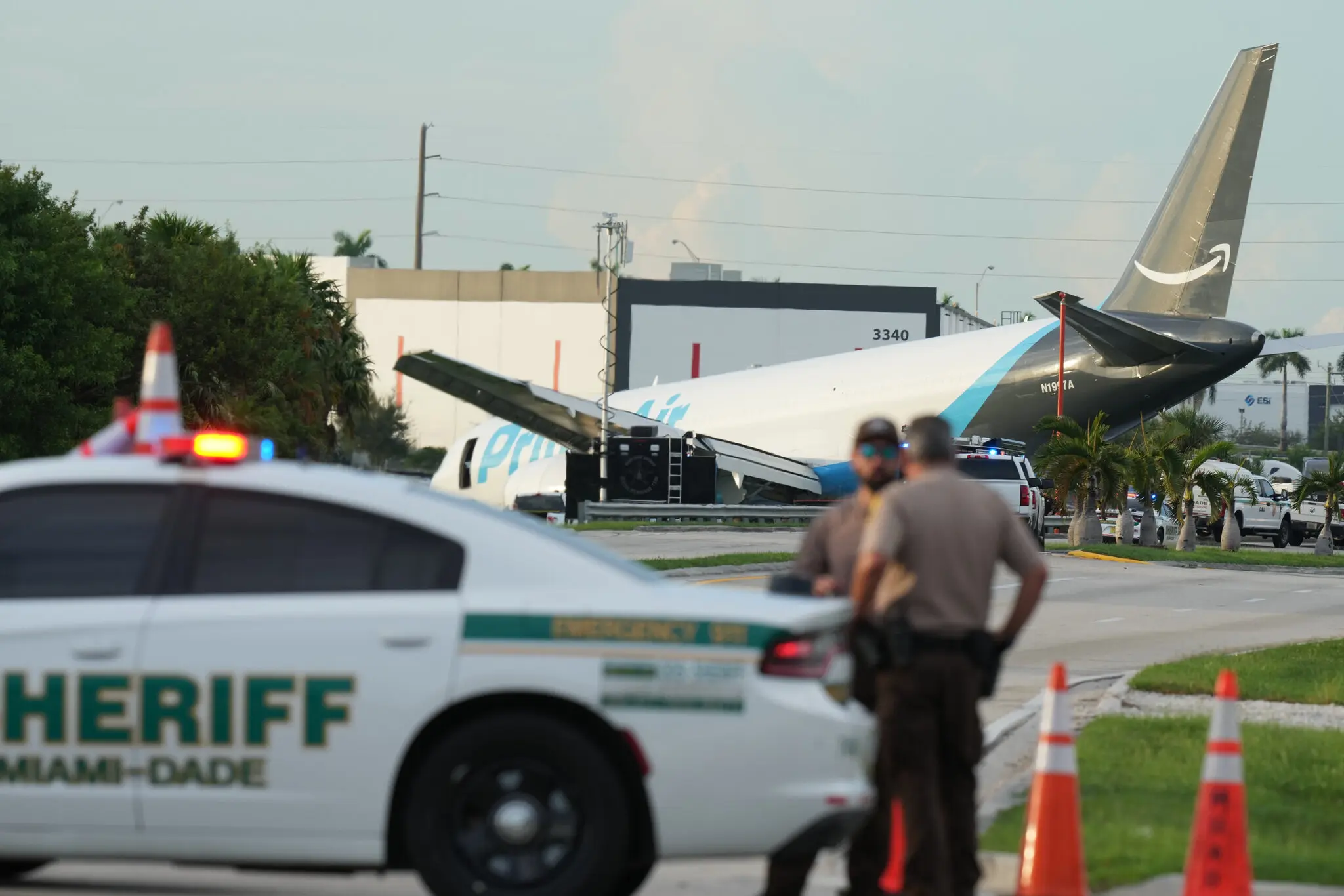 The wreckage of the Amazon cargo plane can be seen with the aircraft's tail elevated in the air. The foreground of the picture shows a Miami police car with two officers stood next to the vehicle.