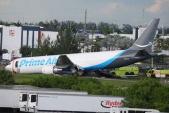 An image taken of the Amazon Cargo plane after the incident. The side of the plane is visibly damaged and emergency response vehicles can be seen in the background.