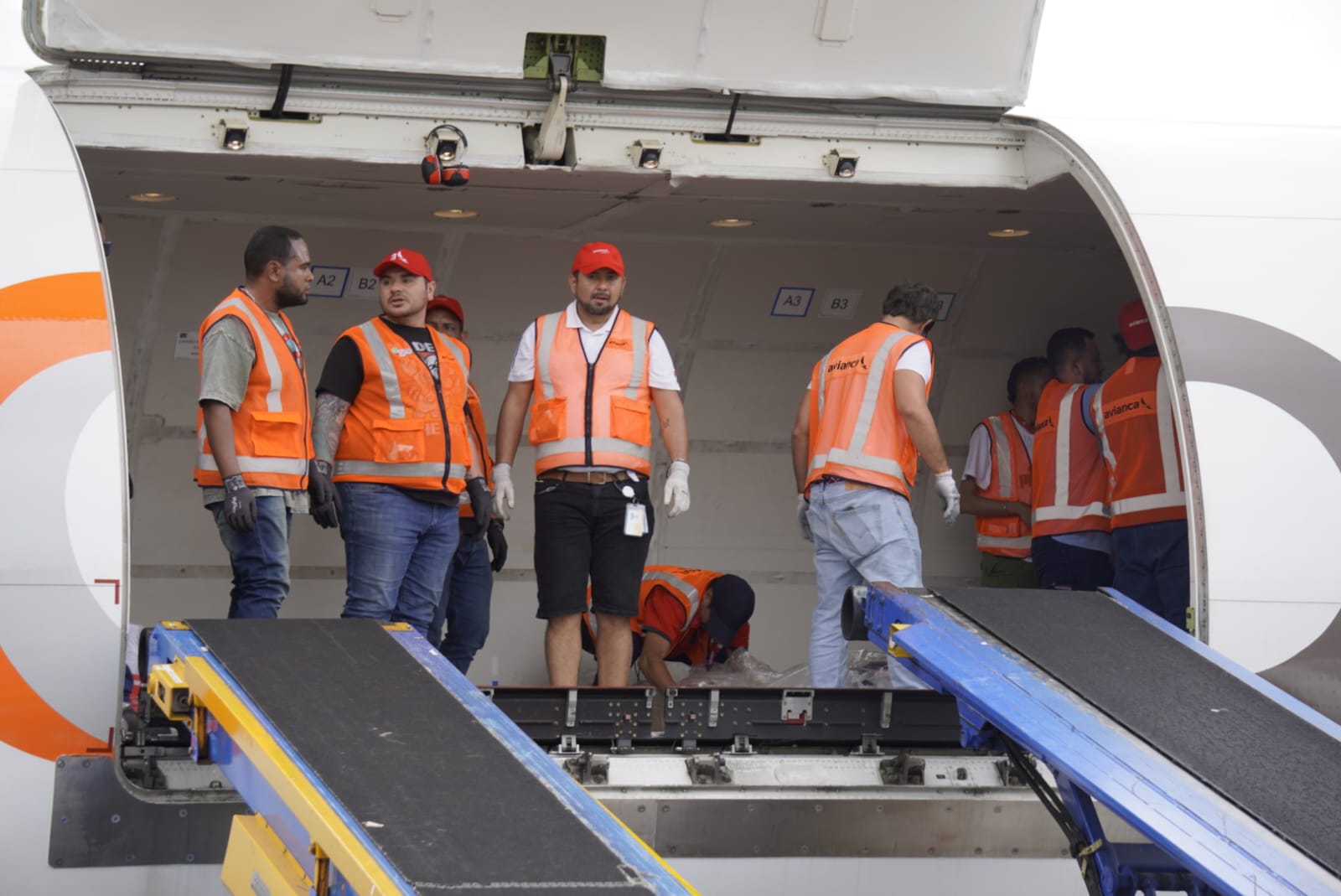 Ground crew workers wearing high-visibility orange safety vests standing inside the open cargo hold of an airplane equipped with conveyor loaders.