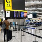 A passenger with a shoulder bag and luggage stands in an airport terminal, looking up at a digital check-in information board near stanchion queue lines.