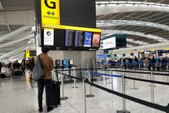 A passenger with a shoulder bag and luggage stands in an airport terminal, looking up at a digital check-in information board near stanchion queue lines.