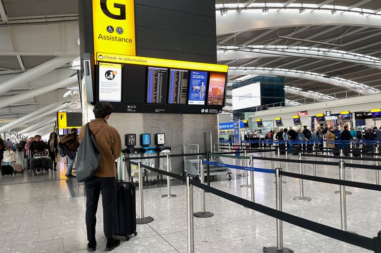 A passenger with a shoulder bag and luggage stands in an airport terminal, looking up at a digital check-in information board near stanchion queue lines.