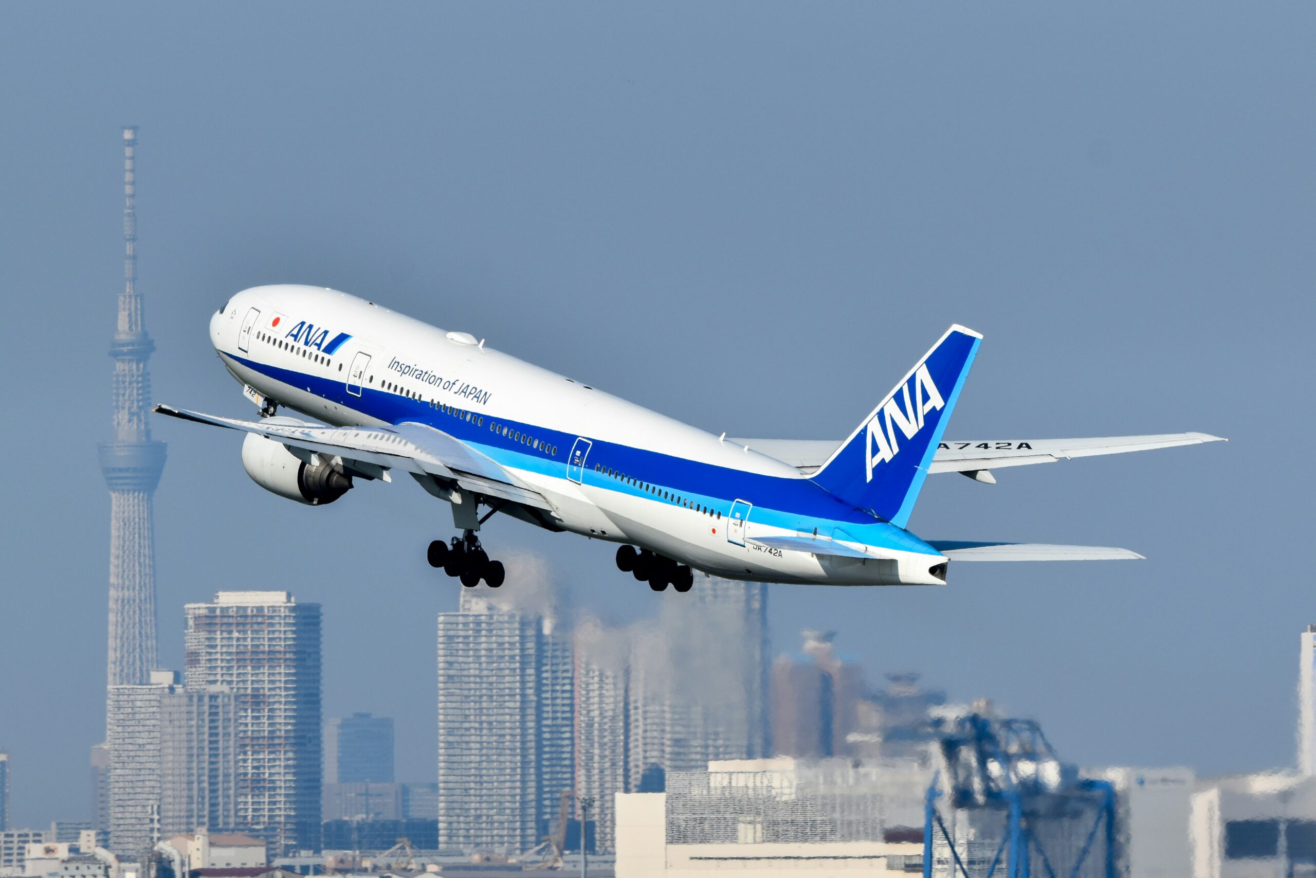 An ANA aircraft midair against skyscrapers in the background. The sky is blue and hazy.