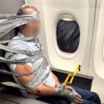 A passenger can be seen physically restrained with duct tape and zip ties. He is bound with several pieces of tape to his seat. His face is blurred.