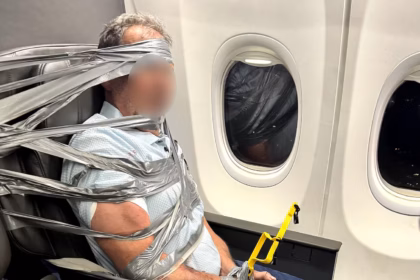 A passenger can be seen physically restrained with duct tape and zip ties. He is bound with several pieces of tape to his seat. His face is blurred.