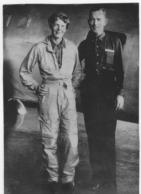 Black and white photo of Earhart and Noonan standing and smiling in front of an aircraft 