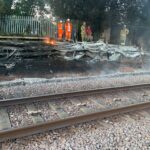 The London Fire Brigade alongside engineers putting out the fire caused by dry grass and heat. They are stood in orange jumpsuits with helmets on to protect them from the fire. There is smoke around the train tracks.