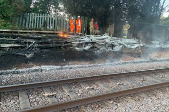 The London Fire Brigade alongside engineers putting out the fire caused by dry grass and heat. They are stood in orange jumpsuits with helmets on to protect them from the fire. There is smoke around the train tracks.