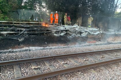 The London Fire Brigade alongside engineers putting out the fire caused by dry grass and heat. They are stood in orange jumpsuits with helmets on to protect them from the fire. There is smoke around the train tracks.