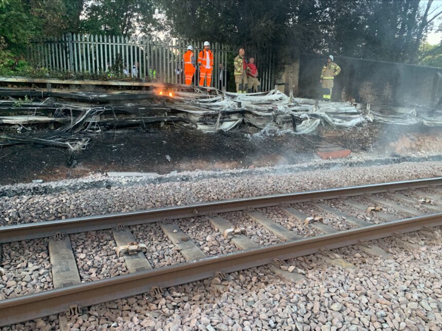 The London Fire Brigade alongside engineers putting out the fire caused by dry grass and heat. They are stood in orange jumpsuits with helmets on to protect them from the fire. There is smoke around the train tracks.