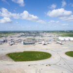 Image shows Heathrow Airport runways in the daytime with grass, aircraft and buildings visible