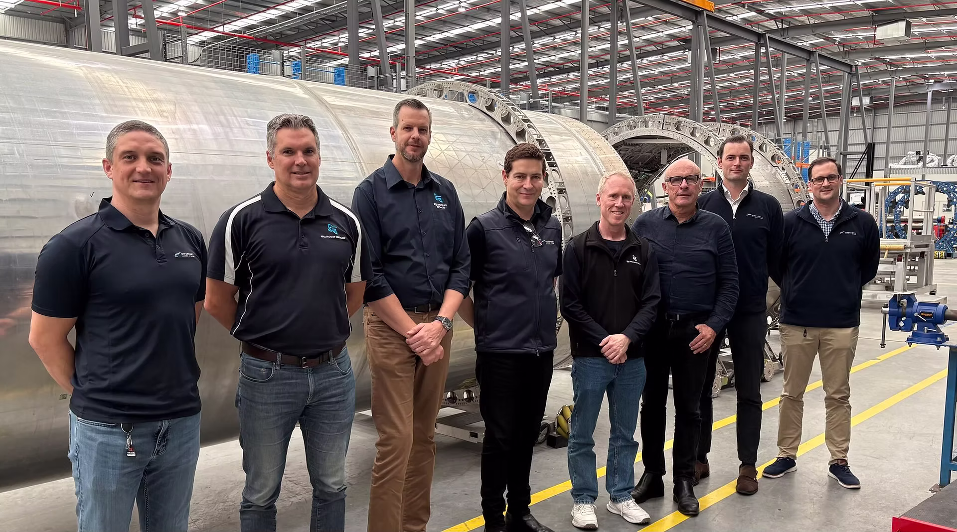 Eight Caucasian males of varying heights and ages stand in a line in what looks like a warehouse or development site. The four men on the left are wearing navy blue Gilmour Space Technologies shirts, and the four on the right are wearing Hypersonix uniform memorabilia. The photograph is taken at an angle, closest to the man pictured on the far right. All of the men face the camera smiling. 