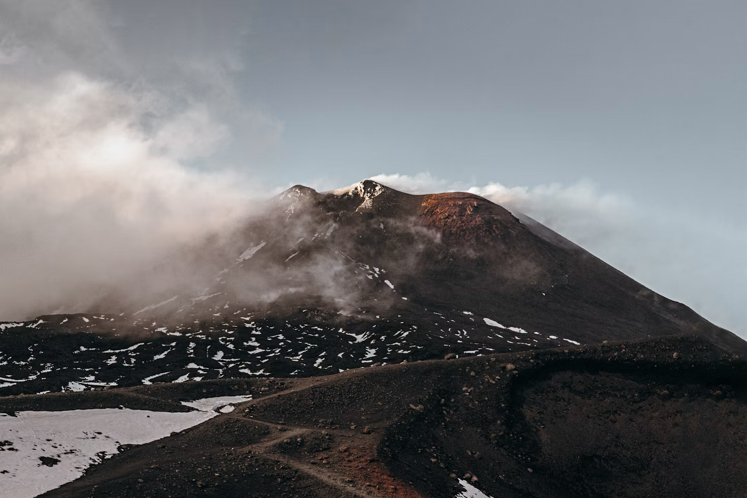 A massive grey volcanic ash cloud over Mount Etna, towering over the mountain. The dark terrain of the volcano stands in front of a bright, clear sky, 