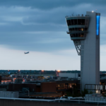 A distant view of a tall, modern air traffic control tower with a unique, angular cab illuminated by a vertical blue light. In the distance, an airplane is flying over an airfield and city skyline at dusk under a cloudy blue sky.