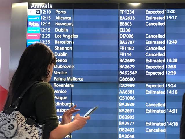 A traveler holding a smartphone stands in profile, looking up at a large digital airport arrivals board showing several delayed and canceled flights.