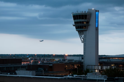 A distant view of a tall, modern air traffic control tower with a unique, angular cab illuminated by a vertical blue light. In the distance, an airplane is flying over an airfield and city skyline at dusk under a cloudy blue sky.