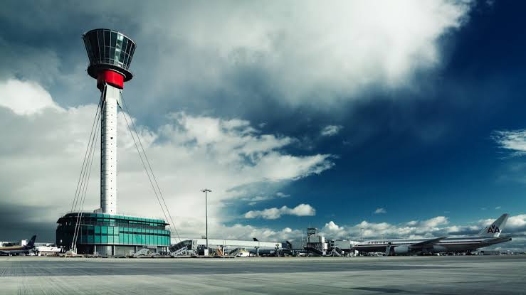 The tall air traffic control tower at London Heathrow Airport with a distinctive red band, standing under a cloudy sky above the tarmac and parked aircraft.