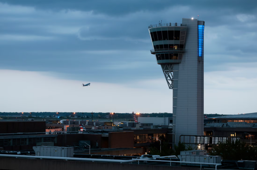 A distant view of a tall, modern air traffic control tower with a unique, angular cab illuminated by a vertical blue light. In the distance, an airplane is flying over an airfield and city skyline at dusk under a cloudy blue sky.