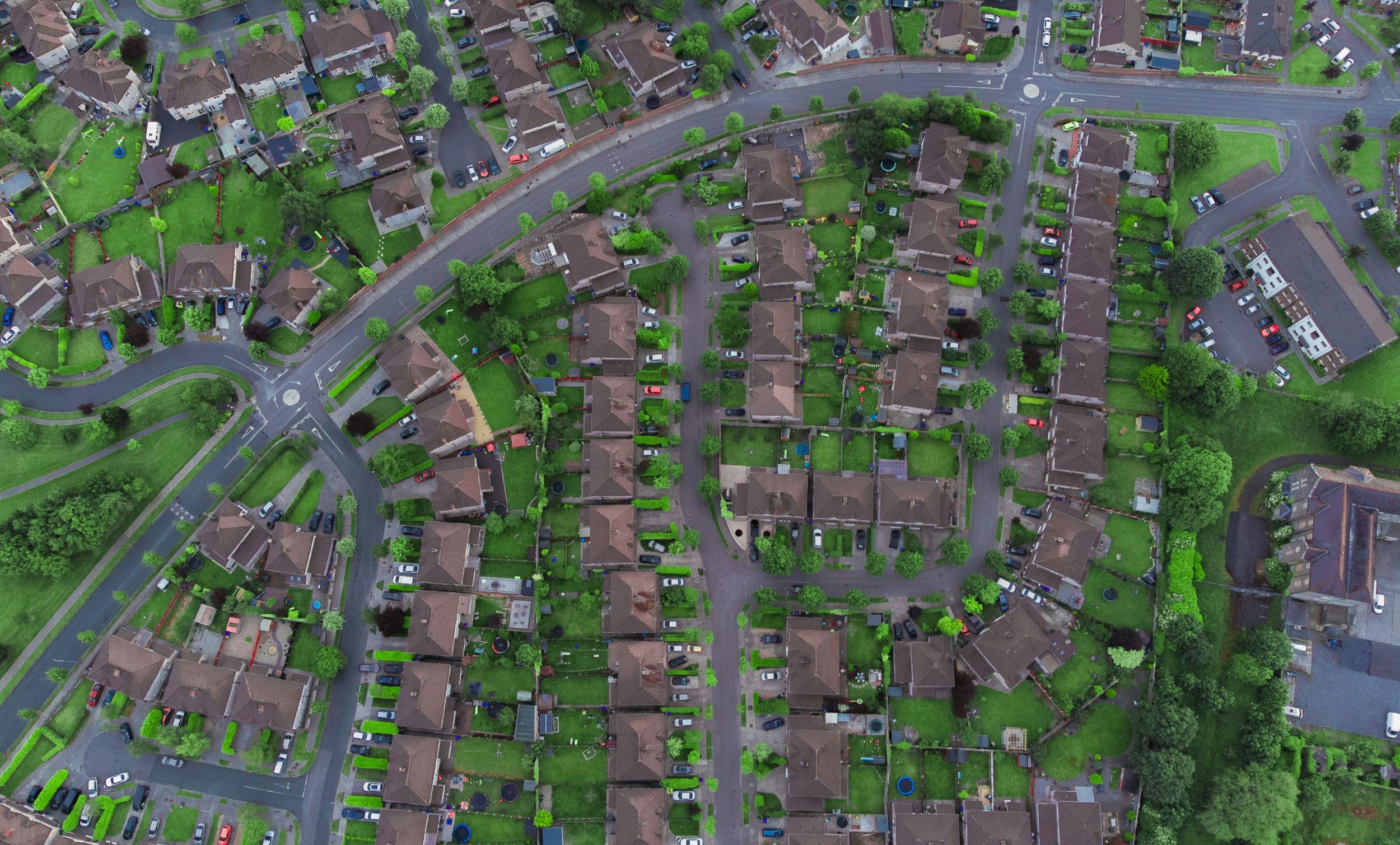 Aerial view of houses in residential area. Brown roofs can be seen across the hole image, interspersed with greenery. A long grey road curves from the bottom left corner up to the top right of the image.