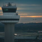 An air control tower at an airport is pictured against a cloudy sky with bright orange light peeking through the clouds. The tower looks out across the airport and beyond hills and trees can be seen.
