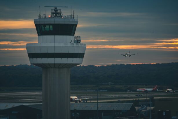 An air control tower at an airport is pictured against a cloudy sky with bright orange light peeking through the clouds. The tower looks out across the airport and beyond hills and trees can be seen.