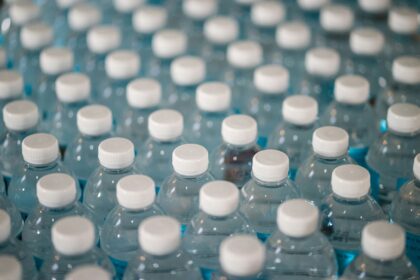 Numerous plastic water bottles are pictured line sup in rows. the image is taken zoomed in/close up from above at slight angle, so they appear in diagonal rows. The bottles become increasingly more stylistically blurred. The bottles are clear with white lids.