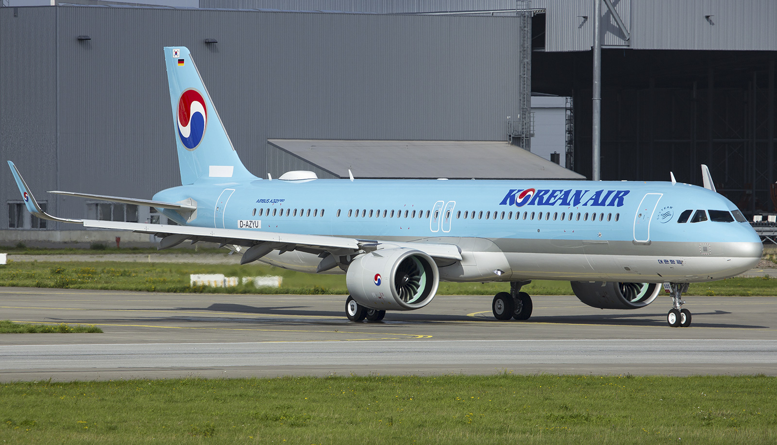 A Korean Air A321neo taxiing on a runway, with an airport building in the background and grass either side of the runway.