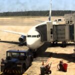 A commercial passenger airplane docked at an airport boarding bridge on a sunny tarmac, surrounded by ground support equipment including a pushback tug vehicle.