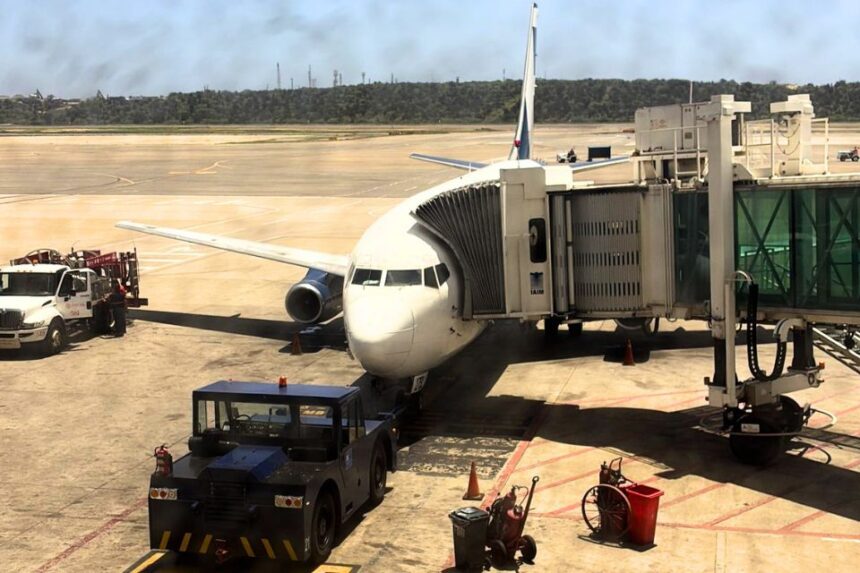 A commercial passenger airplane docked at an airport boarding bridge on a sunny tarmac, surrounded by ground support equipment including a pushback tug vehicle.