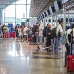 The image shows people in a long queue waiting to be seen at the several security desks at the side of the image.
