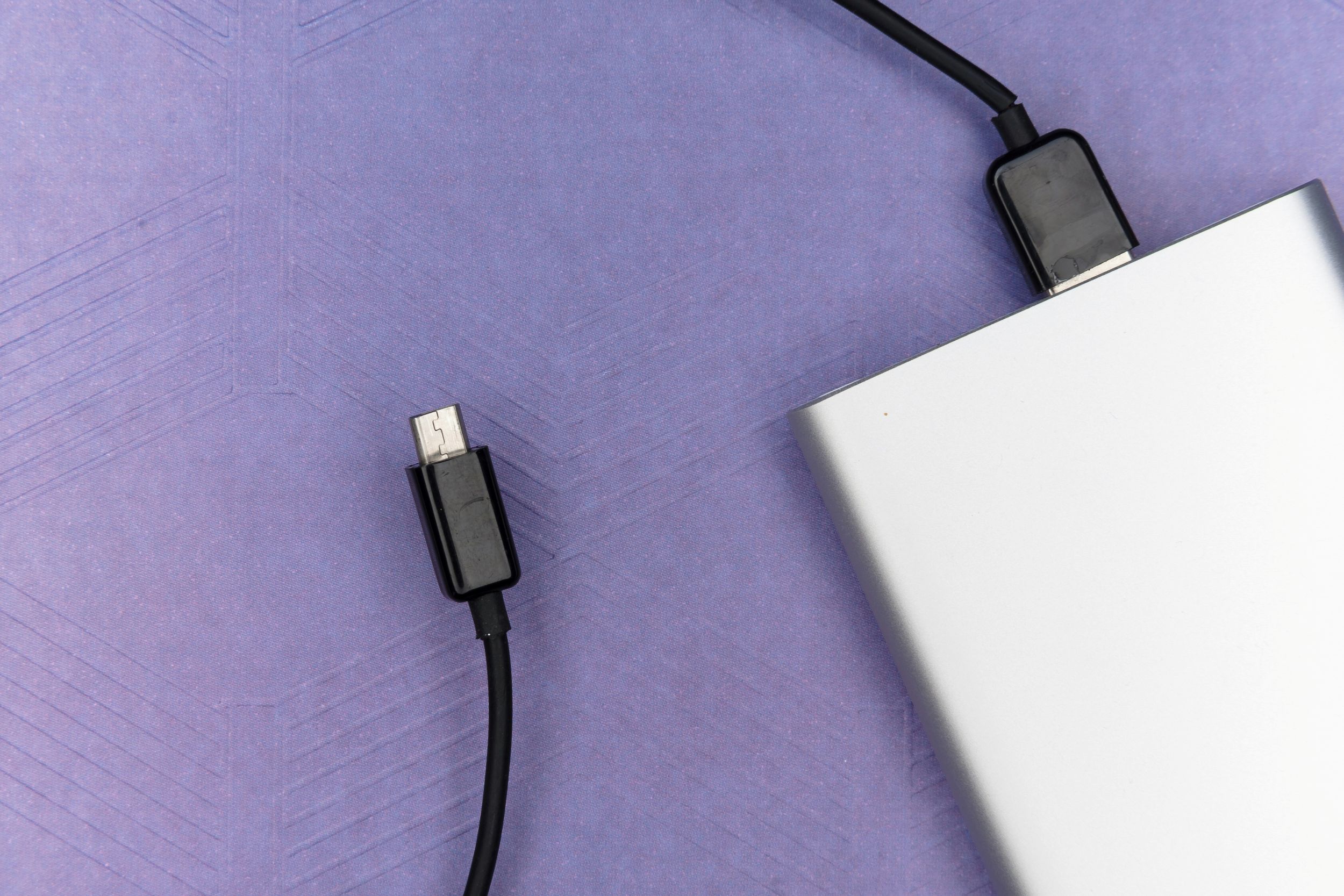 A silver power bank is pictured with a black wire to connect it to a device extending from the top. The power bank is positioned in the bottom right of the image, and the bottom right of the item is not in the picture. The wire extends from the top off screen, with the end re-emerging in the centre right of the picture. The background is a mid purple with faint zig zag texture.