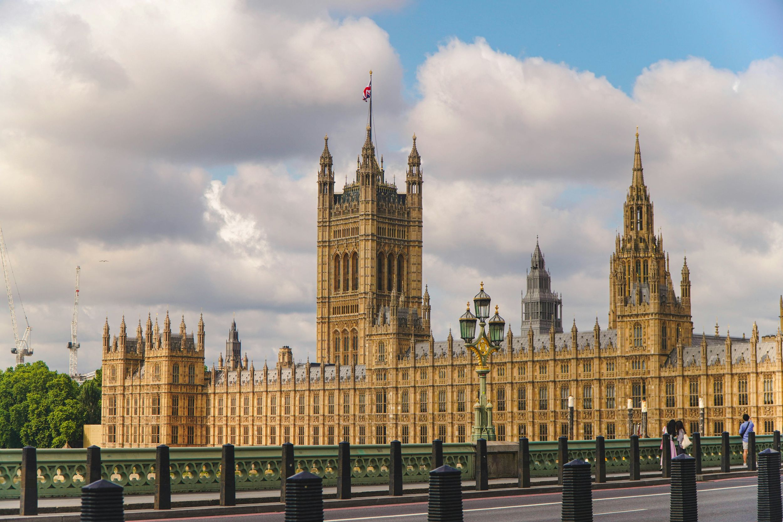 The British Houses of Parliament are pictured from Westminster Bridge during the daytime. The barrier fence that goes along the length of the bridge can be seen in the foreground, with the gothic style tan-yellow parliament building occupying the centre right of the image. The background of the image is a blue sky with lots of large white fluffy clouds.