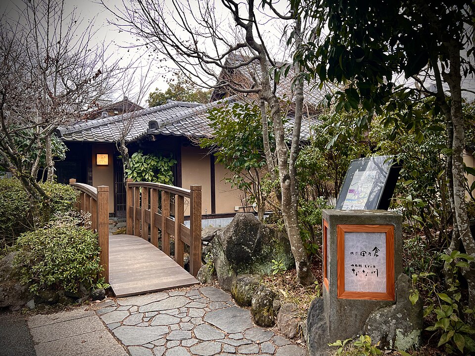 An image of a garden in Oita, Japan. A wooden bridge can be seen leading to a building slightly obscured by trees and foliage.
