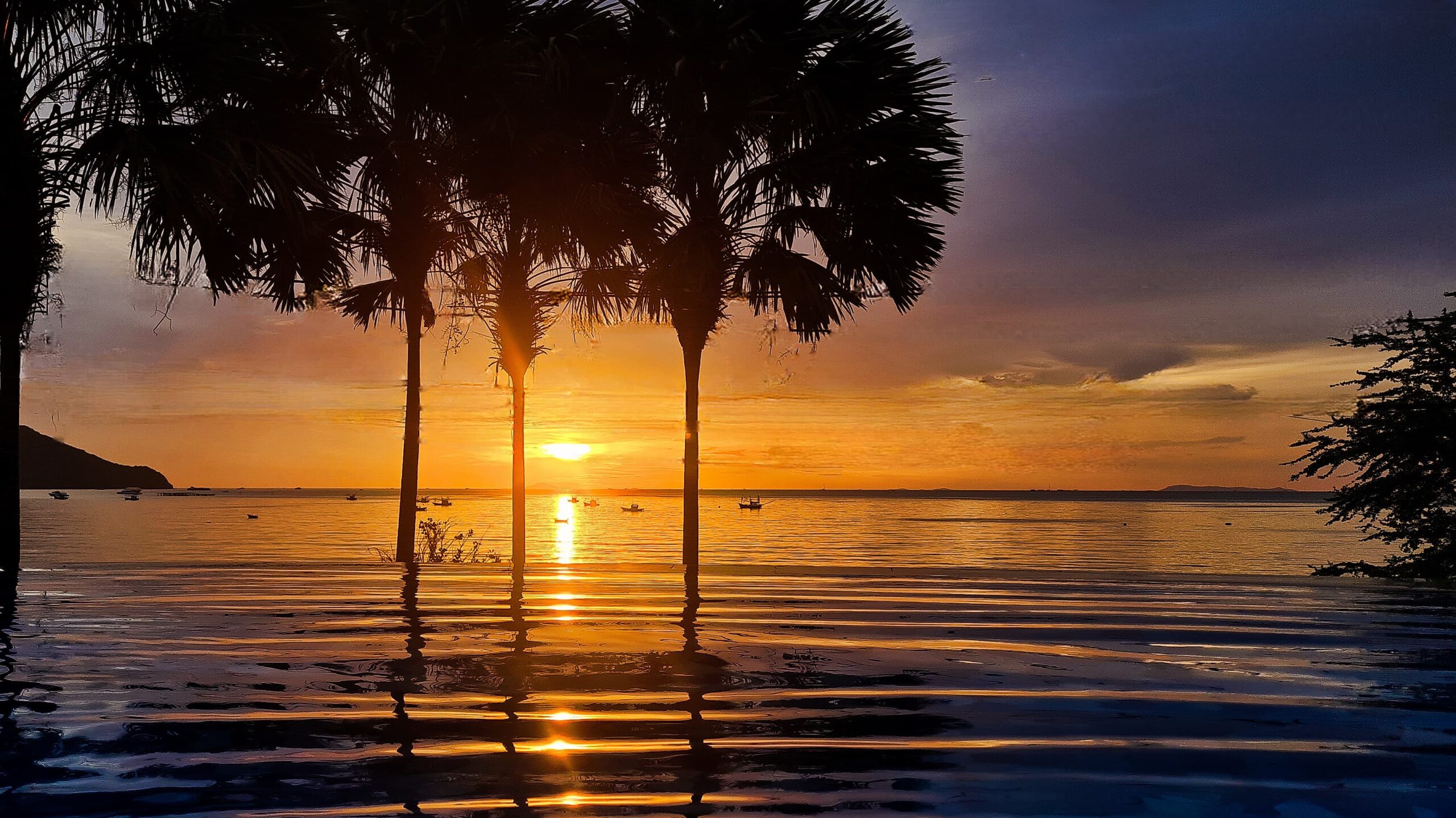 Image shows a sun setting over a calm sea with palm trees and mountains in the background in Thailand