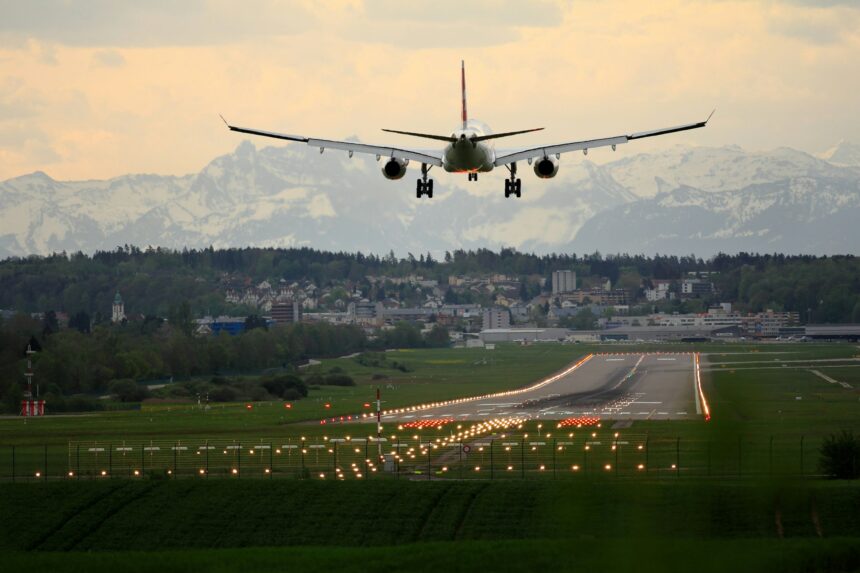An aircraft is approaching a runway to land at either sunrise of sunset. The aircaft is pictured from behind, positioned upper centre right of the image. Either side of the grey tarmac run way is a strip of lights, and grass. In the middle distance, houses can be seen. A golden sky dotted with clouds occupies the background.