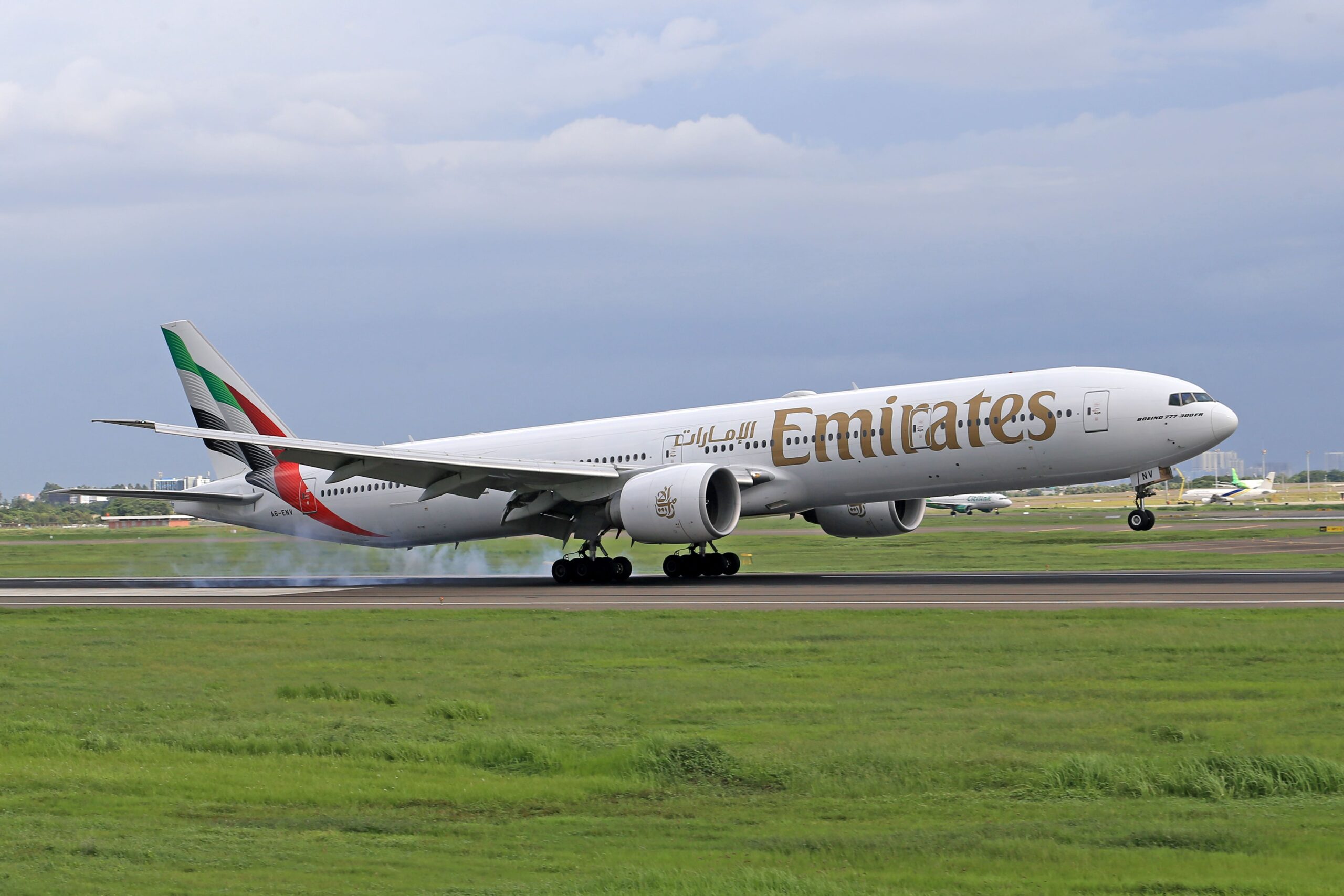 Side view of a white Emirates Boeing 777 commercial airplane landing on an airport runway against an overcast sky. Smoke puffs from the main landing gear as the tires make contact with the tarmac, with grassy fields in the foreground.