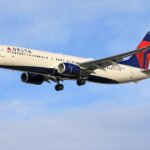 A Delta Air Lines commercial passenger plane, flying under a clear blue sky with its landing gear deployed.