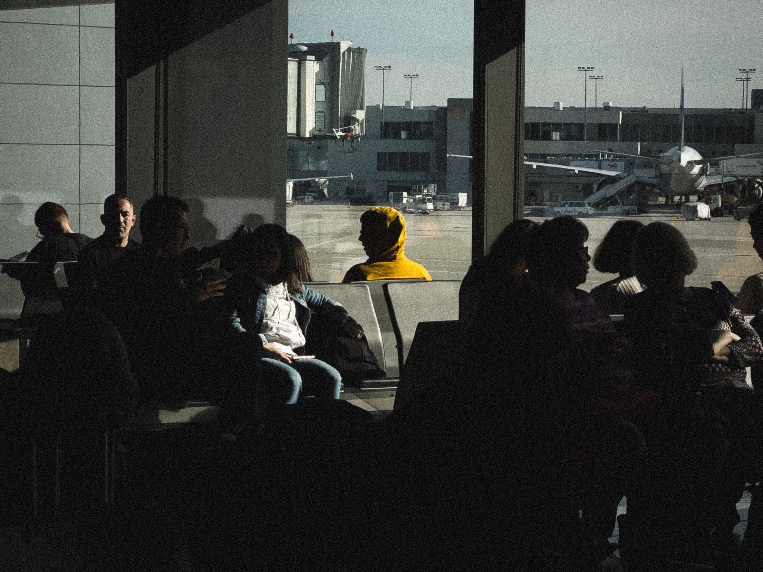Silhouetted travelers in an airport terminal waiting area near large windows with outside tarmac view.