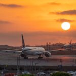 Two wide-body commercial passenger jetliners sit on an airport tarmac during a vivid, glowing orange sunset. The foreground aircraft is viewed from behind as it taxis down the runway, while another plane taxies further in the distance under a bright, low-hanging sun.
