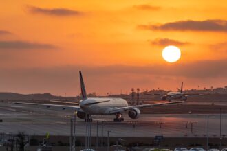 Two wide-body commercial passenger jetliners sit on an airport tarmac during a vivid, glowing orange sunset. The foreground aircraft is viewed from behind as it taxis down the runway, while another plane taxies further in the distance under a bright, low-hanging sun.